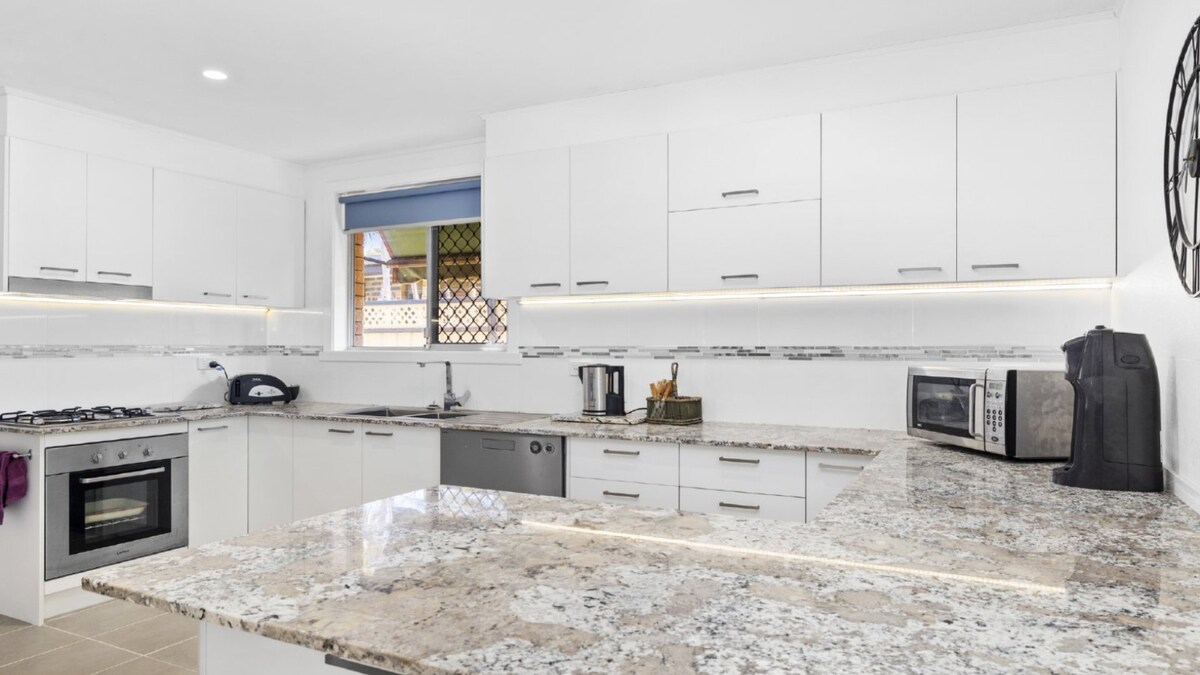 A modern kitchen features crisp white cabinetry and bright lighting. A large granite countertop provides ample workspace, while stainless steel appliances, including a microwave and dishwasher, are neatly arranged. Natural light filters in through a window above the sink, creating a bright atmosphere.