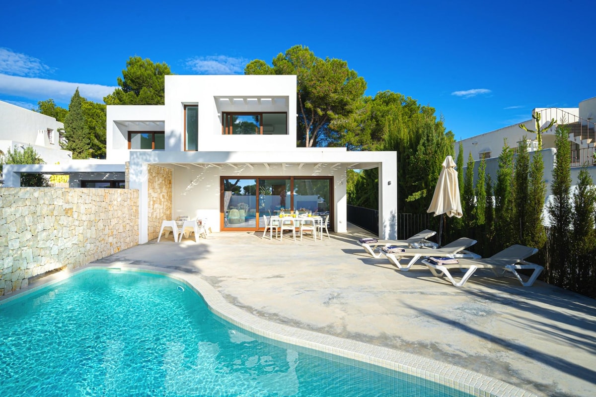 A modern villa is displayed, featuring a private pool and spacious sun terrace. Lounge chairs are arranged nearby, and a dining area is visible under a covered patio. The exterior showcases a blend of contemporary materials and surrounding greenery, offering a serene atmosphere.