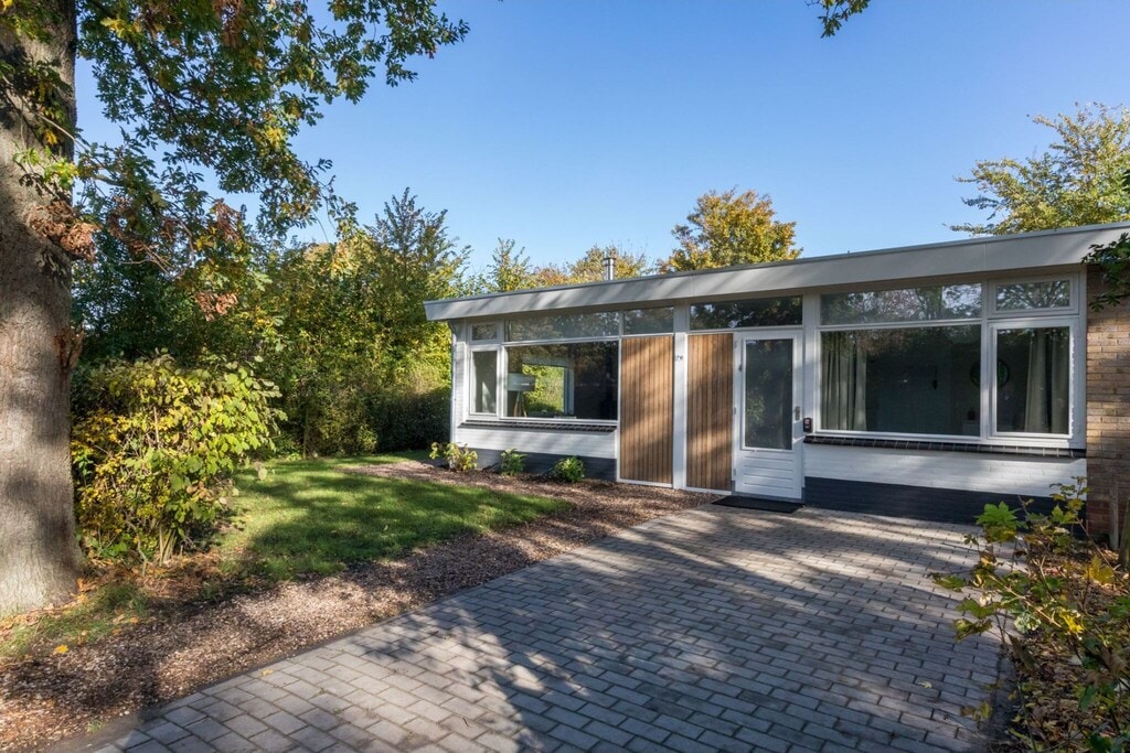 The exterior of a modern house is showcased, featuring large windows that allow natural light to fill the interior. A spacious pathway of cobblestones leads to the entrance, surrounded by a well-maintained garden with greenery and seasonal foliage.