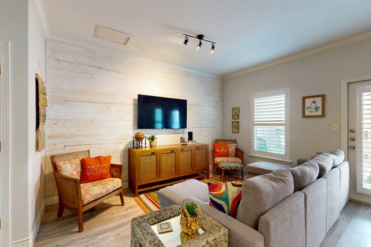 A comfortable living area showcases a sectional sofa arranged around a vibrant area rug. A wooden console houses a flatscreen TV mounted above it. Two cozy chairs are positioned nearby, with decorative cushions adding color. Natural light filters through the window, enhancing the inviting space.