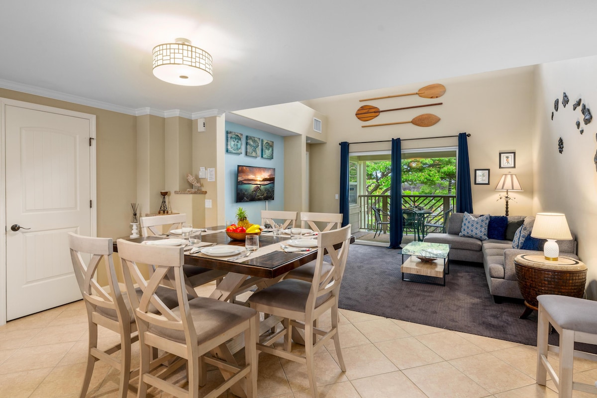 An open-concept living space is presented, featuring a dining table set for six. A cozy seating area includes a sofa and side table, with natural light streaming through glass doors that lead to a private balcony surrounded by greenery.