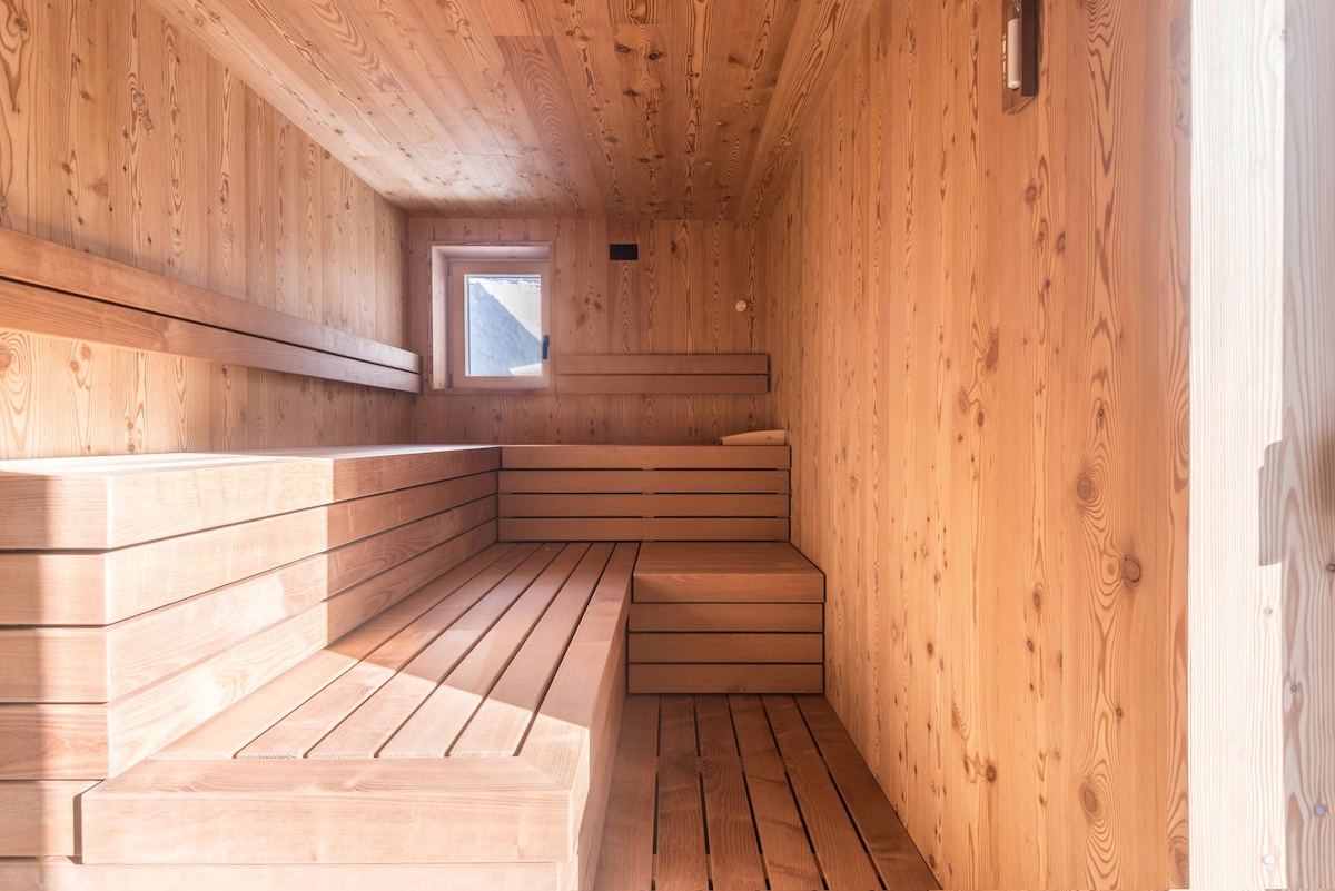 A wooden sauna space is shown with slatted benches lining the walls. Natural light filters in through a window, enhancing the warm timber tones of the interior. The minimalistic design emphasizes a tranquil environment for relaxation.