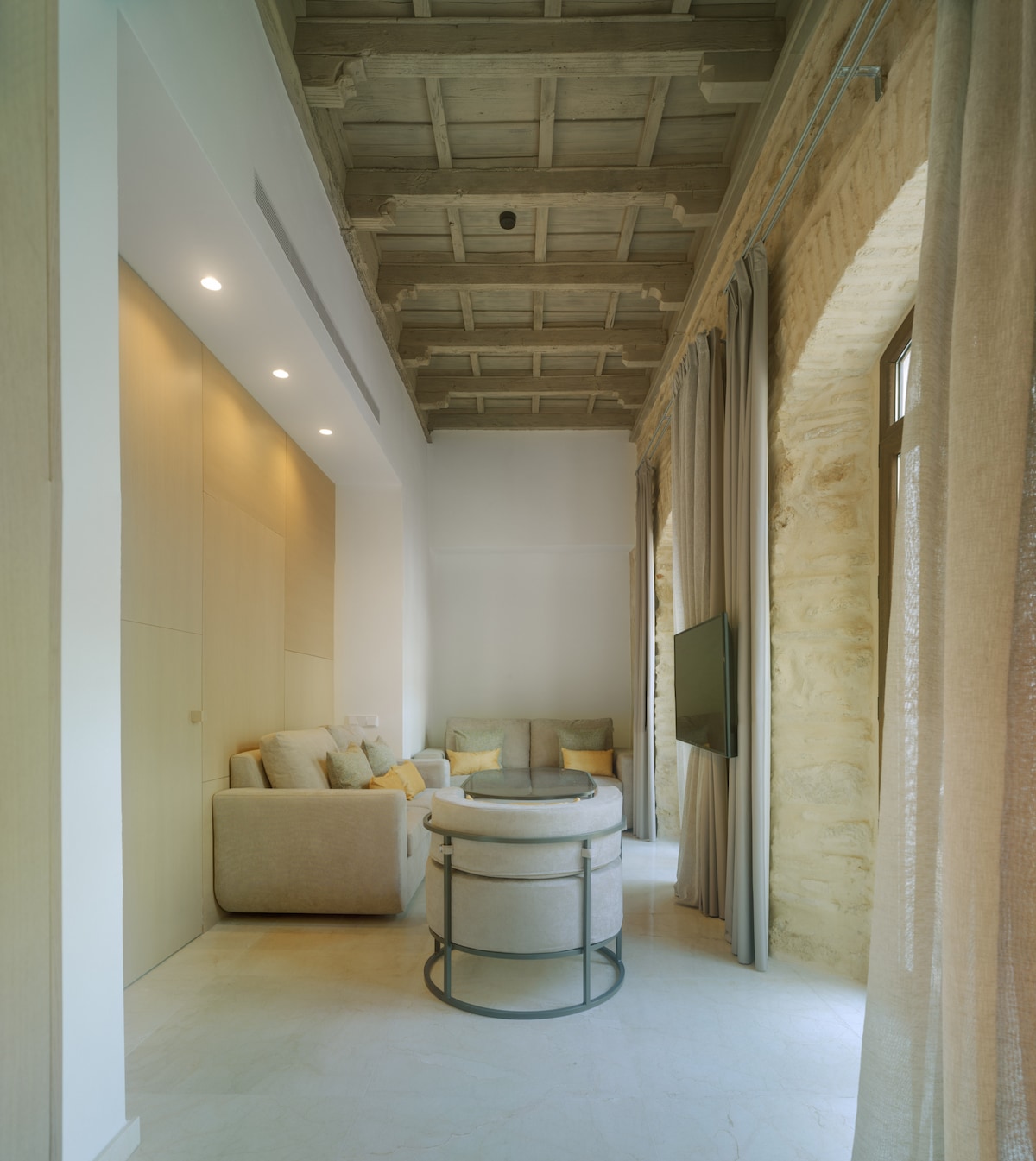 The living area features a relaxed seating arrangement with two light-colored sofas. A circular coffee table is placed in the center, complemented by soft cushions in muted tones. Exposed wooden beams enhance the ceiling, while large windows allow natural light to fill the space.