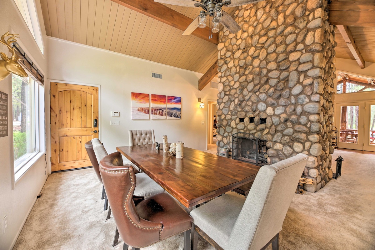 A spacious dining area features a wooden table surrounded by a mix of upholstered and wooden chairs. A distinctive stone fireplace serves as a focal point against the light-colored walls, with natural light flooding the room from large windows.