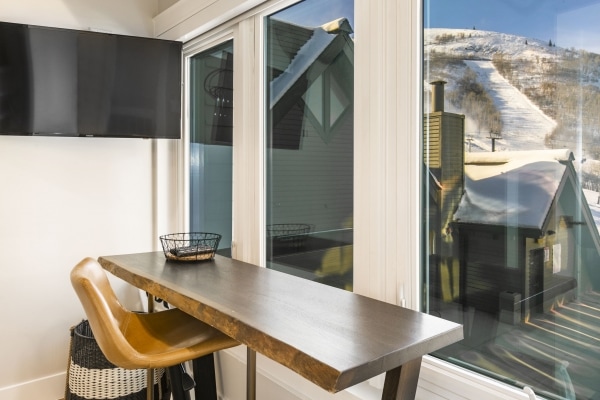 A sleek workspace is positioned next to large windows, offering views of snowy hills. A simple wooden table is complemented by a modern chair, while a small decorative basket adds a touch of elegance. A flat-screen TV is mounted on the wall nearby.