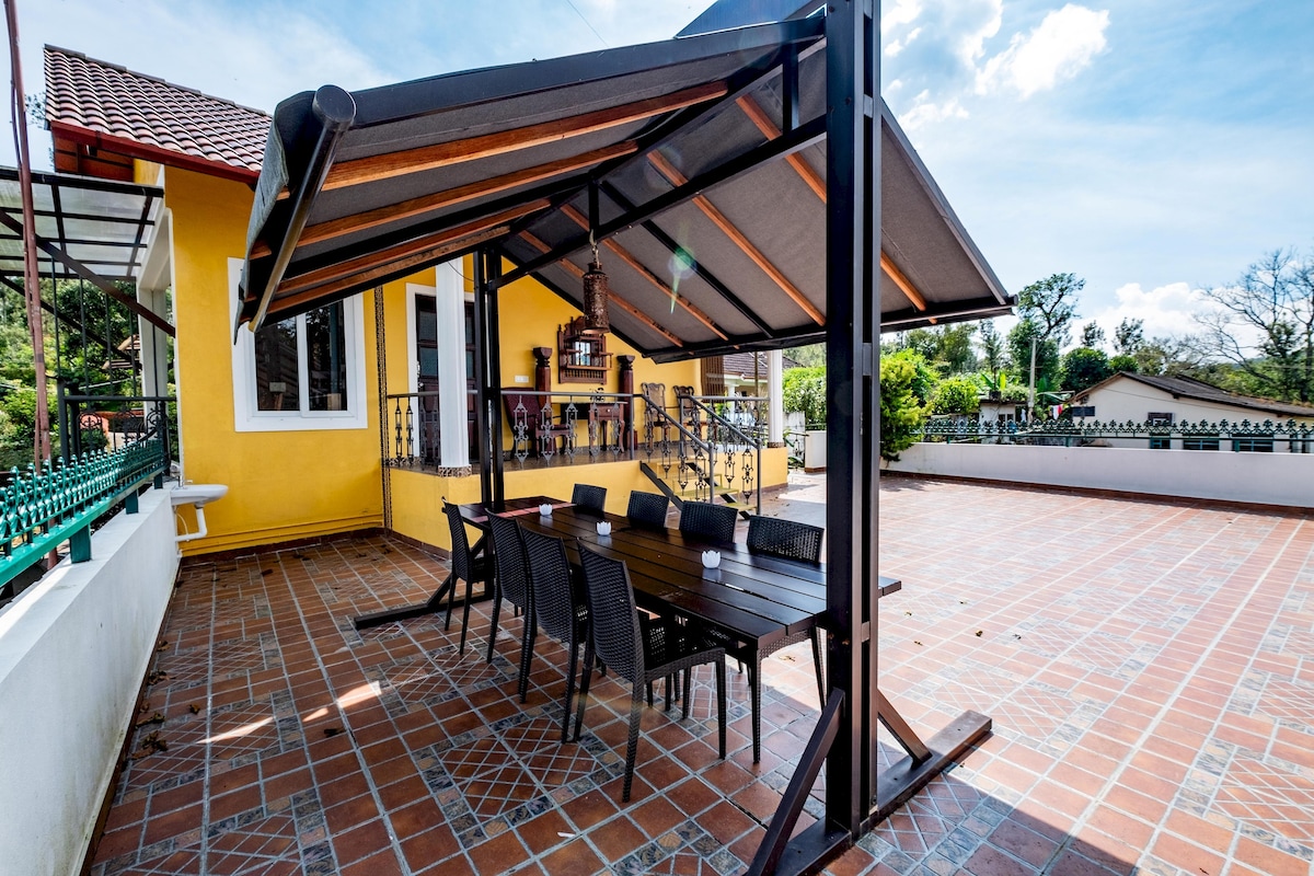 An outdoor shaded dining area is featured, with a large table surrounded by several chairs. The space is situated on a tiled patio, adjacent to a yellow house with a balcony. Green foliage and distant trees provide a natural backdrop.