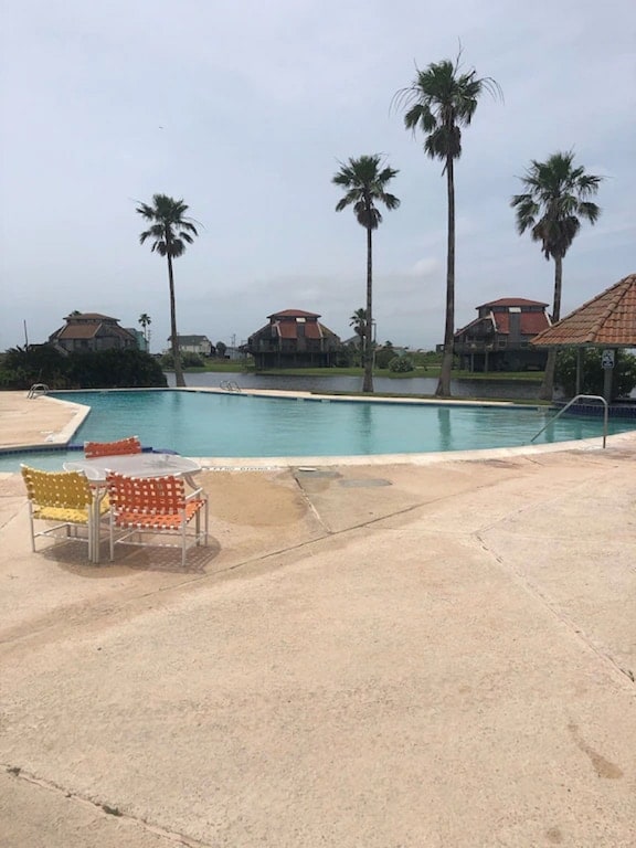 A serene pool area is framed by palm trees, with a spacious swimming pool reflecting the sky. Colorful lounge chairs in orange and yellow are arranged around the poolside, inviting relaxation. Scenic views of nearby structures can be seen in the background.