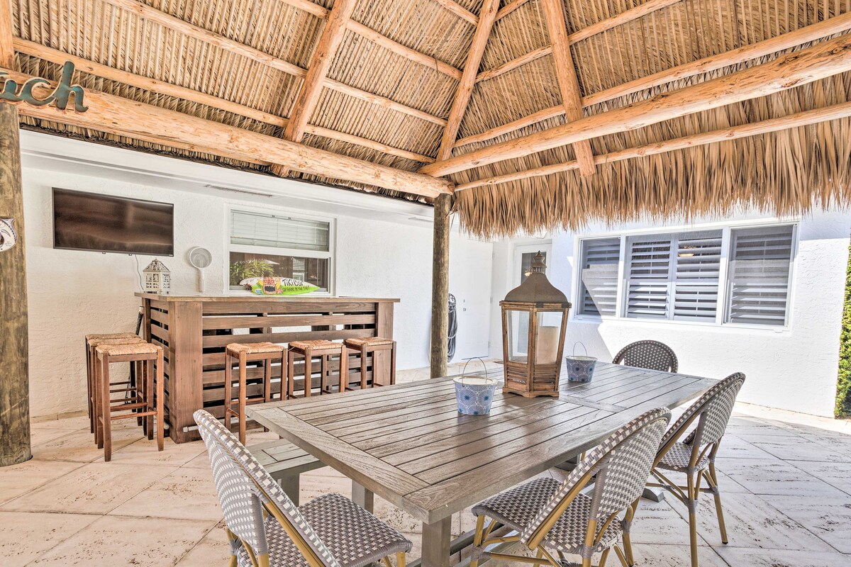 A spacious outdoor tiki hut is highlighted with a thatched roof, featuring a large wooden dining table and matching bar seating. A gas lantern and decorative elements add charm to the inviting space. Nearby, windows allow natural light to brighten the area.