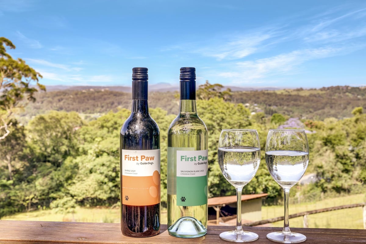 Two bottles of wine labeled 'First Paw' are positioned on a wooden table, accompanied by two glasses filled with water. In the background, lush greenery and rolling hills are visible under a clear blue sky.