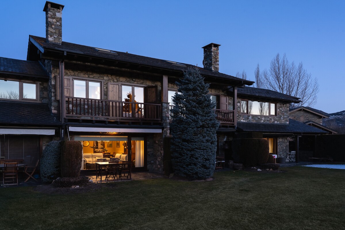 The exterior of the rustic three-story home is highlighted by stone accents, showcasing a balcony on the upper level. Warm light spills from the windows, creating a welcoming glow against the twilight backdrop. Lush greenery surrounds the property, providing a cozy outdoor space.
