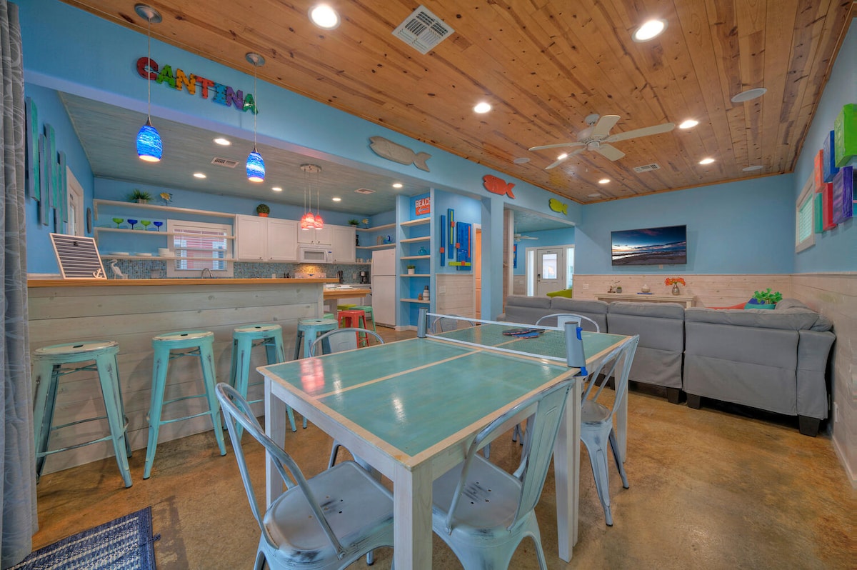 A bright and airy dining area features a large table with turquoise accents surrounded by white chairs. The walls are painted in a cheerful blue, complemented by colorful artwork. A comfortable seating area with gray sofas is visible in the background.