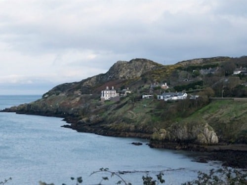 A scenic coastal view captures a rocky shoreline complemented by gentle waves. A cluster of charming homes is nestled against the hillside, framed by lush greenery and distant rolling hills. The sky displays a mix of clouds and blue, contributing to the serene ambiance.
