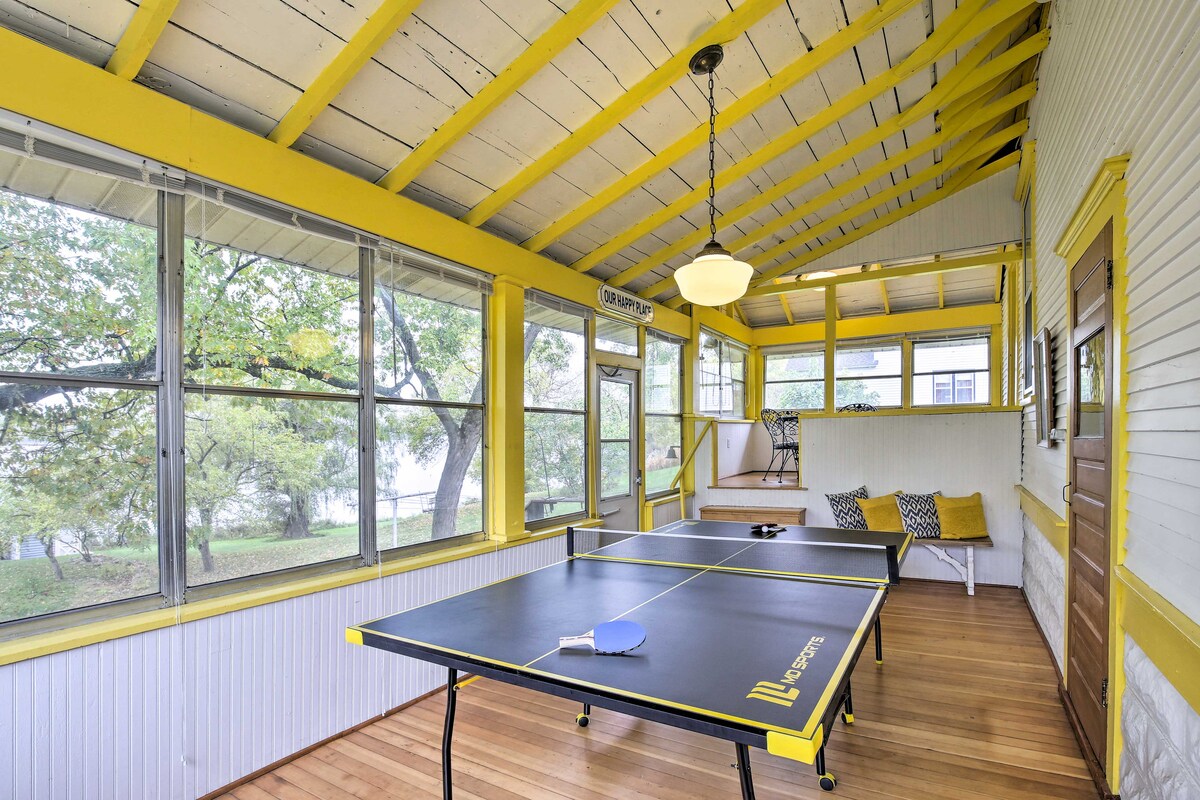 The sunroom features a ping pong table positioned centrally on hardwood flooring. Large windows allow natural light to illuminate the space, adorned with yellow beams and white walls. A cozy seating area is visible in the corner, enhancing the room's inviting atmosphere.