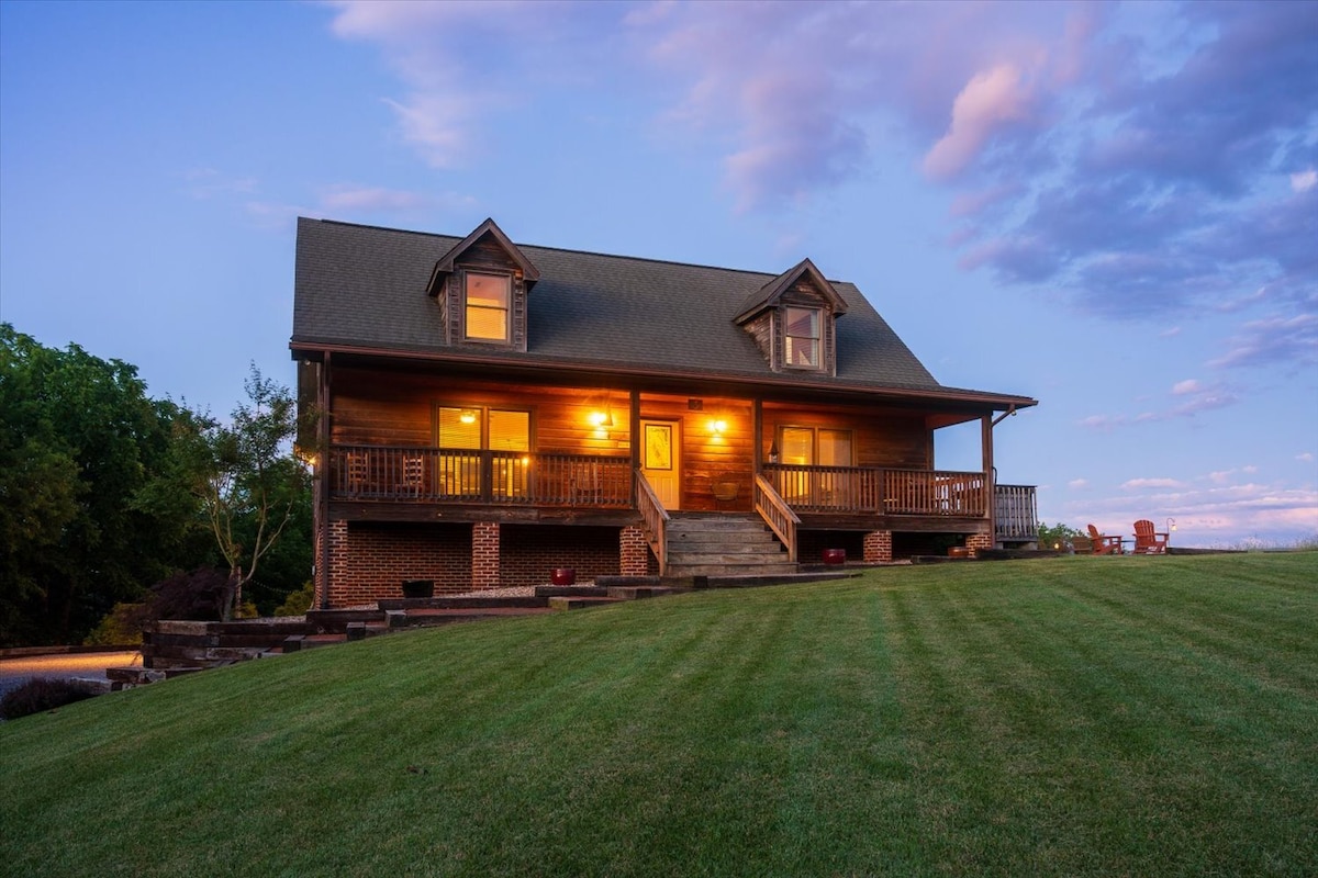 The cabin exterior features a welcoming porch with steps leading to a landscaped yard. Soft evening light casts a warm glow from the windows, highlighting the charming architecture against a backdrop of colorful twilight sky. The expansive green lawn extends towards the horizon.