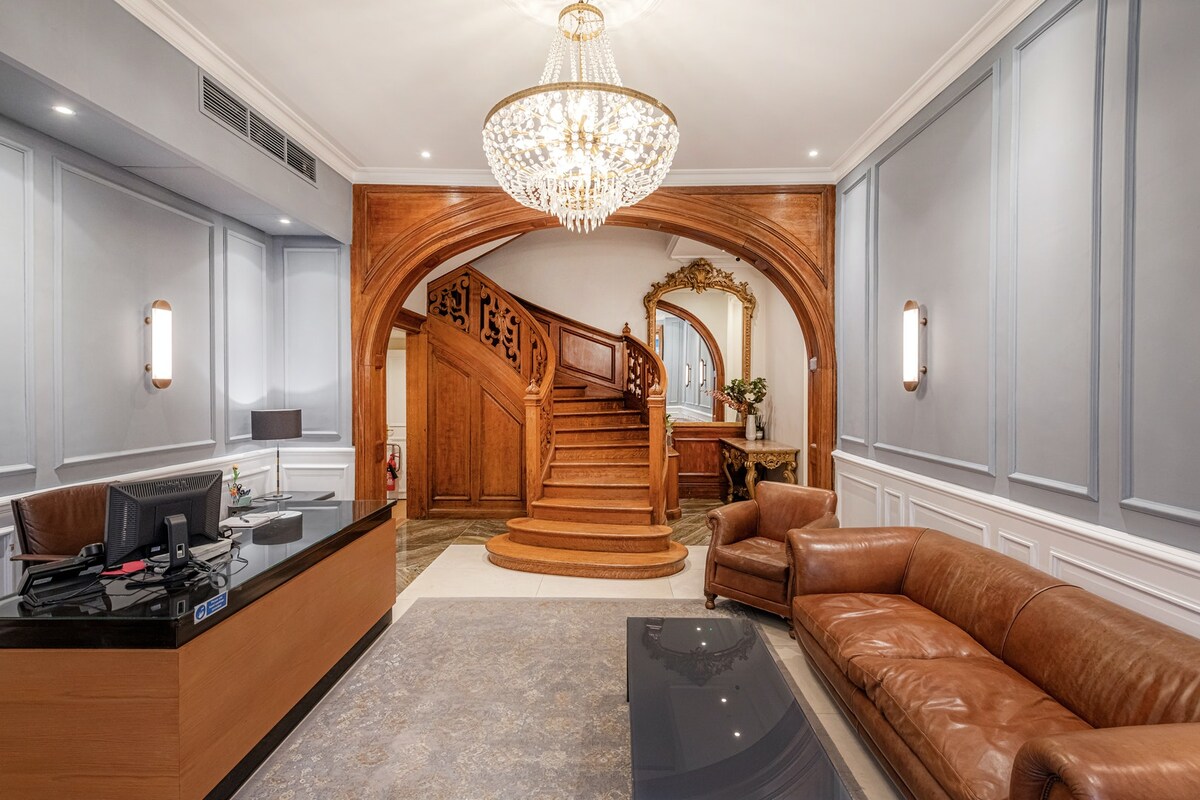 A welcoming reception area features elegant woodwork and a chandelier that enhances the space. A reception desk is situated on one side, with a plush brown sofa and coffee table providing comfortable seating. The grand staircase is prominently displayed, adding to the overall sophistication.