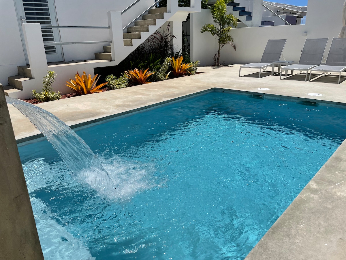 A private pool area features crystal-clear water with a gentle fountain stream. Surrounding the pool are four chaise lounge chairs positioned for sunbathing, alongside vibrant tropical plants and landscaping that enhance the tranquil outdoor space.