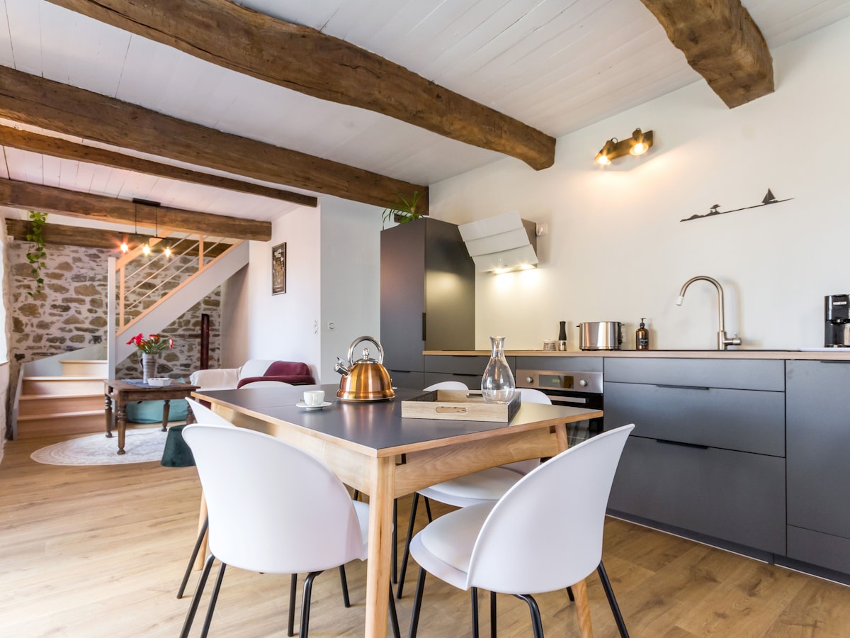 An open kitchen is presented with modern appliances and a wooden dining table surrounded by four white chairs. Exposed wooden beams adorn the ceiling, adding character to the space. A staircase leads to the upper floor, while natural light brightens the area.