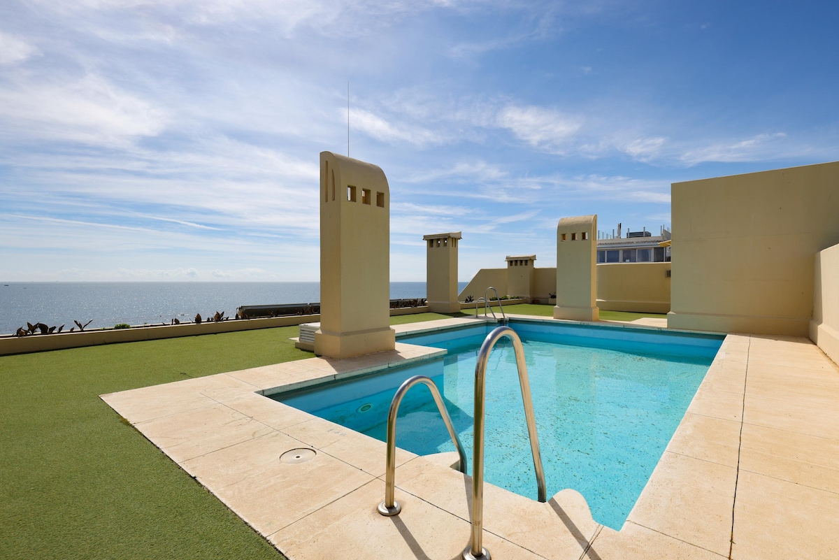 An outdoor pool is surrounded by a green lawn, offering views of the sea and clear blue sky. The pool area is designed with a minimalist aesthetic, featuring a light-colored stone deck and steps leading down into the water.