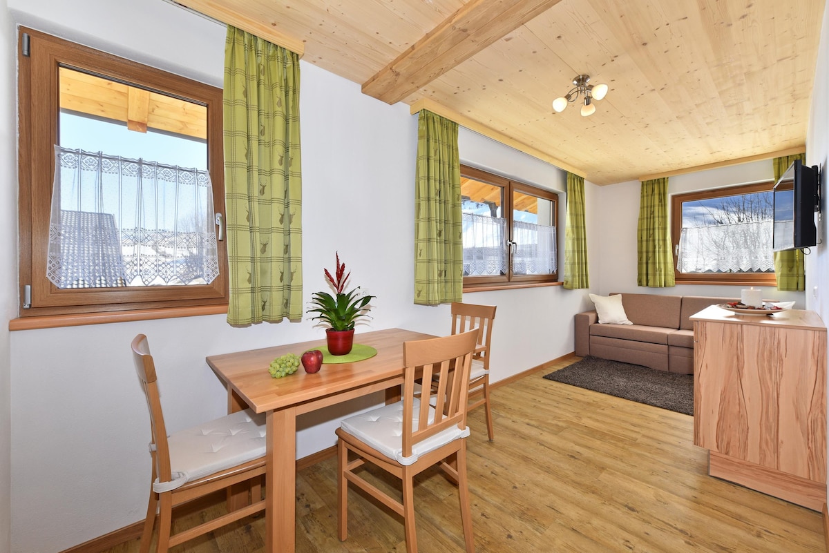 The living area features natural wood accents and light-colored walls. A small dining table with two chairs is complemented by a cozy sofa. Large windows with green curtains allow natural light to fill the space, offering views of the outdoors.