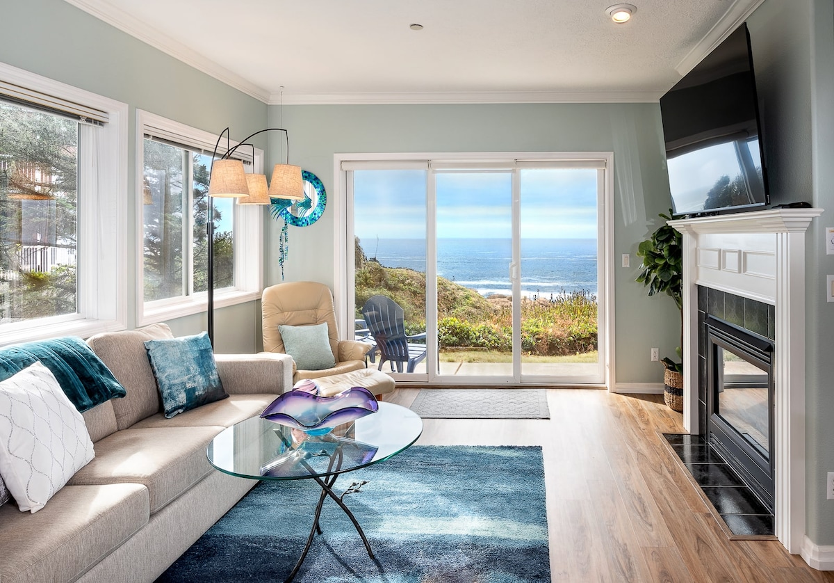 The living area features a light-colored sofa and a glass coffee table atop a soft area rug. Natural light floods in through large windows with ocean views, highlighting the gas fireplace beside a mounted flat-screen TV. A comfortable chair and outdoor seating are available on the balcony.