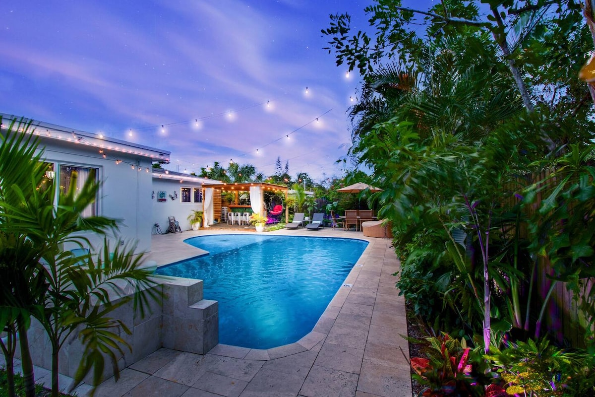 A serene outdoor area showcases a heated saltwater pool, surrounded by lush greenery and vibrant plants. Illuminating string lights hang above, creating a calm atmosphere. Comfortable lounge chairs and an outdoor dining area are visible, inviting relaxation and socialization.