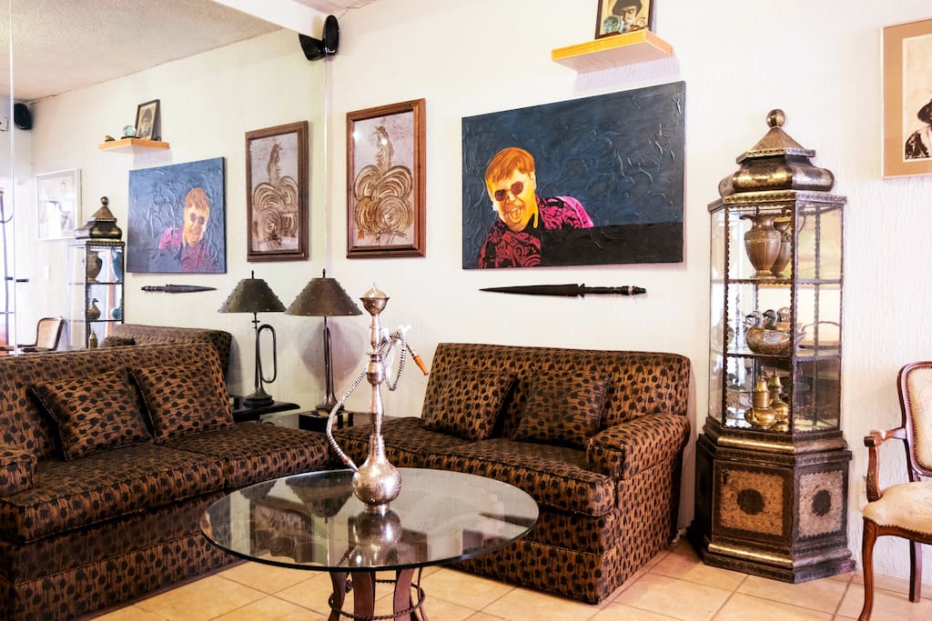 A cozy living area features two patterned sofas and a glass coffee table at its center. Artwork adorns the walls, creating an engaging focal point. Unique decor items, including lamps and a glass display case, contribute to the eclectic design, enhancing the room's character.