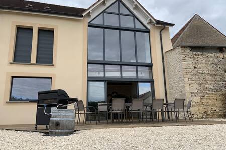 The exterior of the villa is depicted, featuring large glass windows that enhance natural light. A spacious deck is furnished with multiple chairs around a dining table, with a barbecue grill nearby. The structure is complemented by a stone wall, blending contemporary and traditional styles.