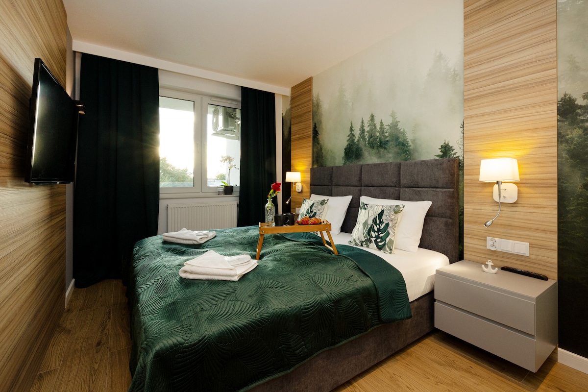 The bedroom features a spacious bed covered with a green quilt and decorative pillows. A minimalistic bedside table and a TV are present, while a large window is adorned with dark curtains. Subtle wall art depicts a serene forest scene, enhancing the calming ambiance.
