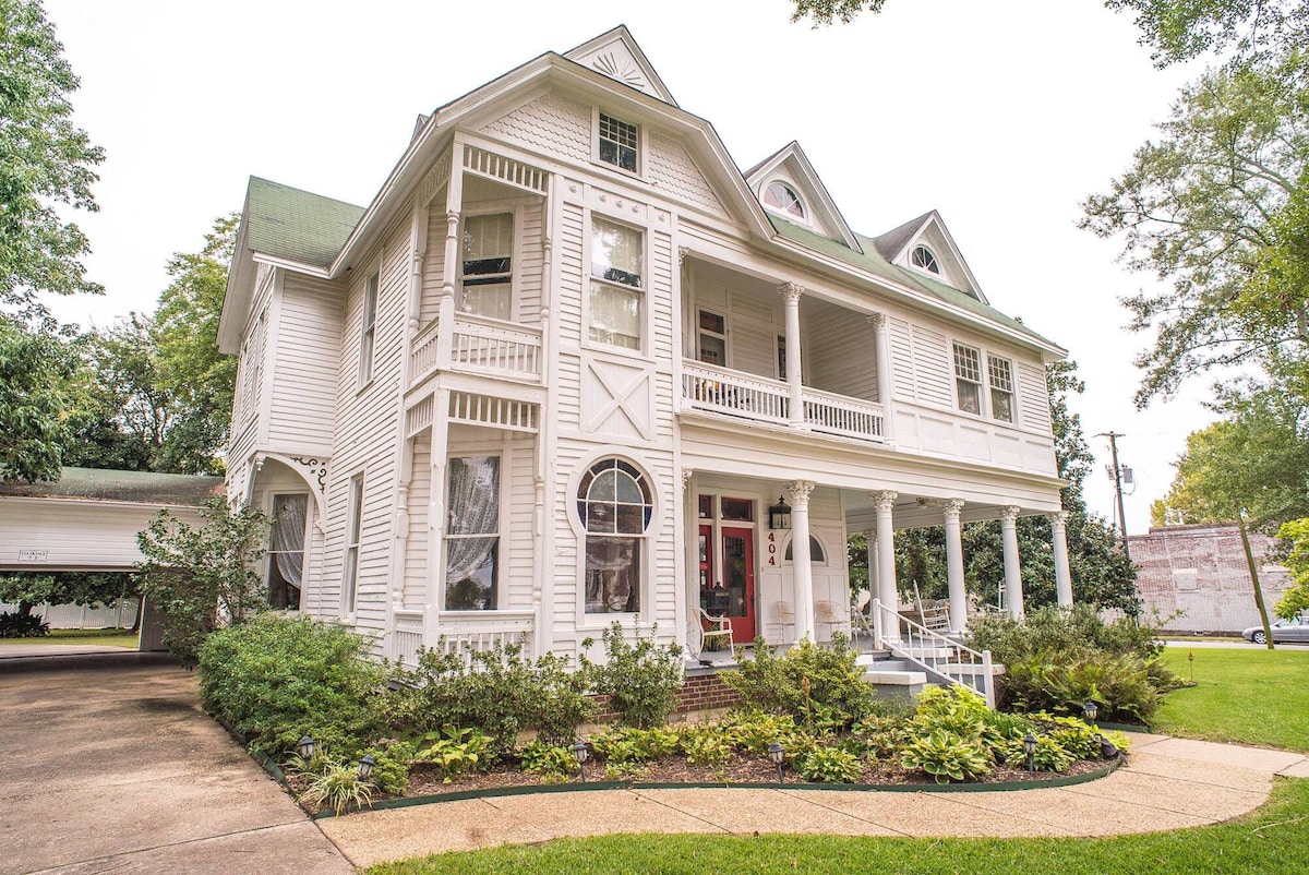 A charming two-story white house is surrounded by lush greenery. The exterior features a welcoming porch with several steps leading to the entrance. Large windows allow natural light into the interior. A curved pathway guides guests through the well-kept landscaping.