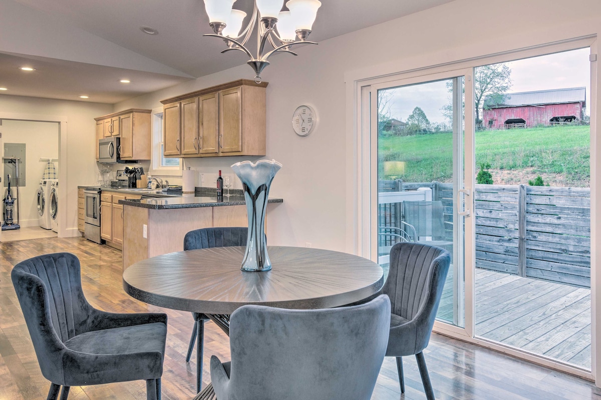 A dining area features a round table surrounded by plush chairs, with a modern chandelier overhead. Sliding glass doors open to the outdoor deck, revealing a view of the greenery beyond. The kitchen area is visible in the background, equipped with stainless steel appliances.