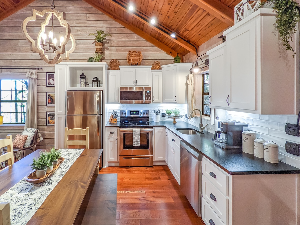 The kitchen features modern stainless steel appliances integrated into a rustic wood interior. White cabinetry contrasts with dark countertops. A central island provides additional workspace and seating. Natural light floods the space through windows, enhancing the cozy atmosphere created by wooden beams overhead.