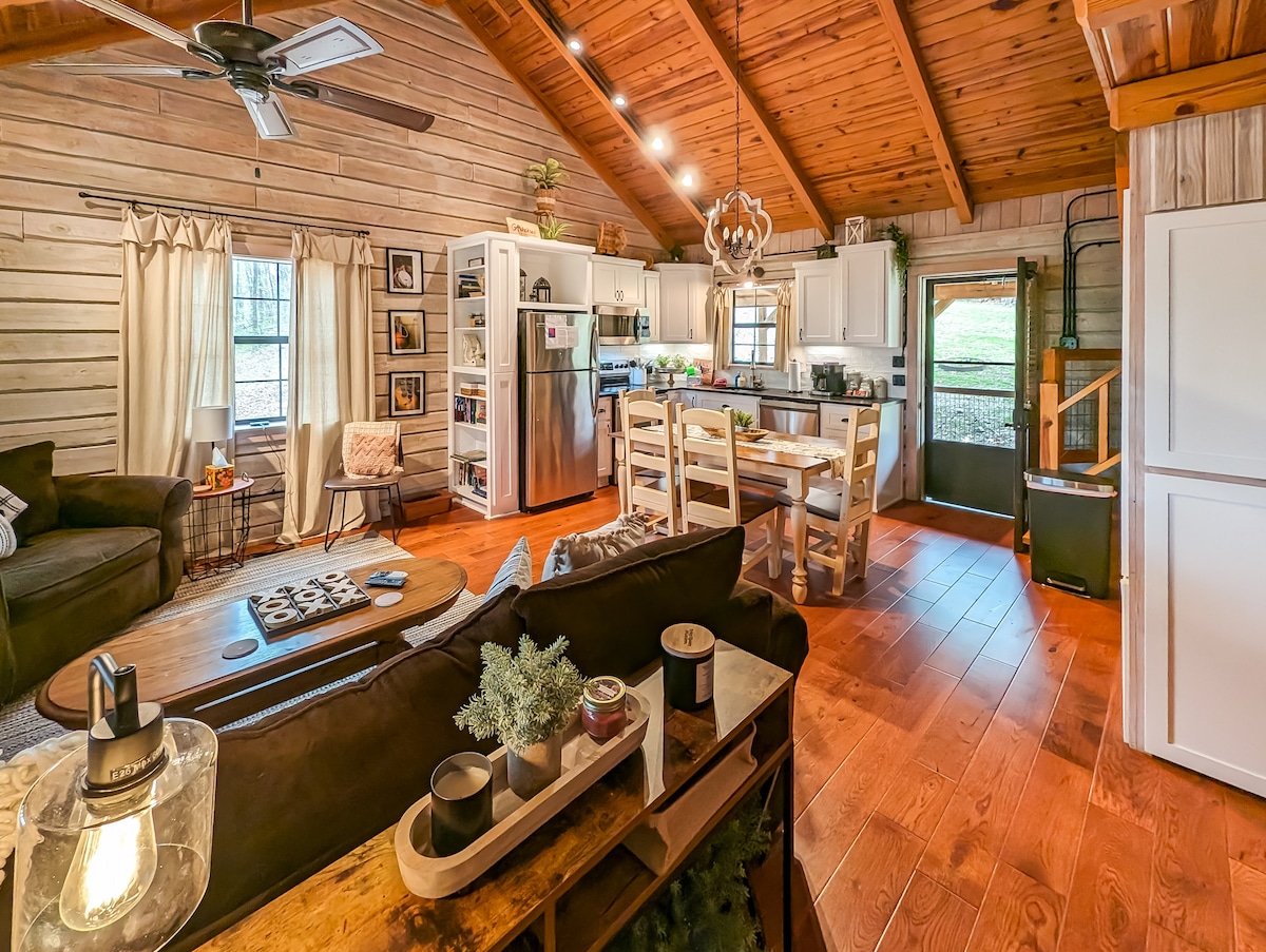 The open living area features a cozy seating arrangement with a dark sofa, complemented by warm wooden walls and a high ceiling. A dining table is positioned adjacent to a well-equipped kitchen, while natural light fills the space through windows, enhancing the inviting atmosphere.