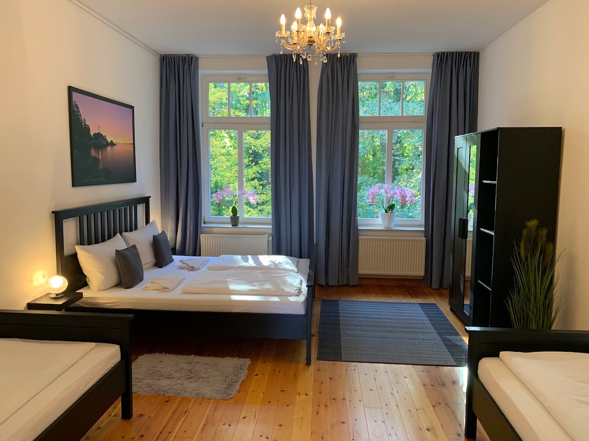A well-furnished bedroom features a spacious bed with crisp linens and an additional bed. Large windows allow natural light to illuminate the room, complemented by elegant curtains. A tasteful piece of artwork hangs above the bed, and a wardrobe stands against the wall.