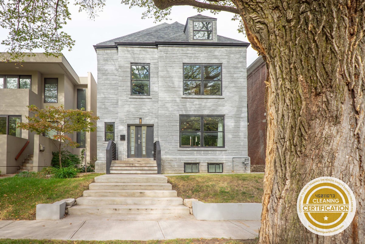 The exterior of a spacious grey home is shown, featuring a classic design with large windows. A set of wide stairs leads up to the main entrance, framed by a large green tree. The surrounding area is well-maintained and inviting.