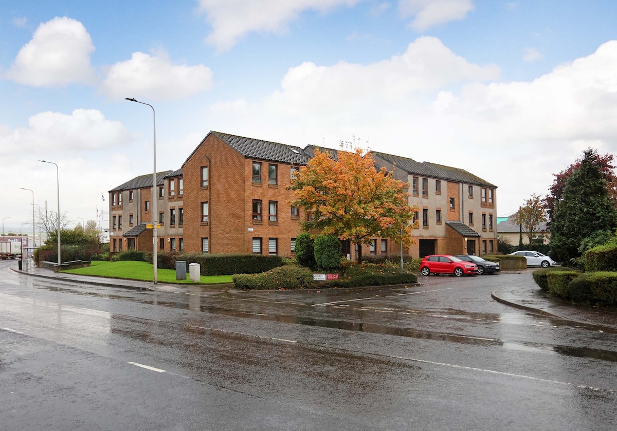 A modern, two-story brick building is situated in a residential area, surrounded by neatly maintained lawns and trees. The scene is set on a rainy day, with wet pavement reflecting the cloudy sky. A red car is parked nearby.