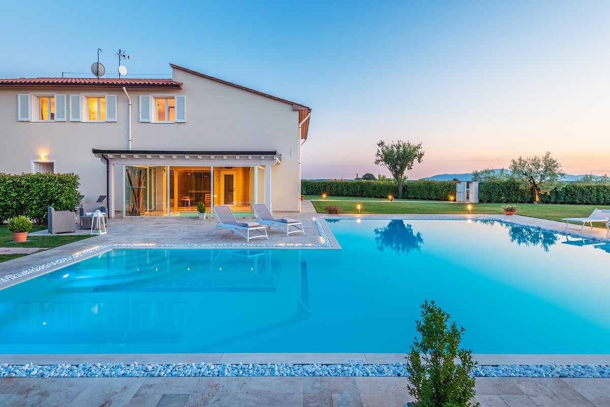 The villa features a large private swimming pool, surrounded by a tiled area and well-maintained garden. Sun loungers are positioned near the pool, offering relaxed seating options. The two-story structure provides a clear reflection in the water, complementing the serene outdoor setting.