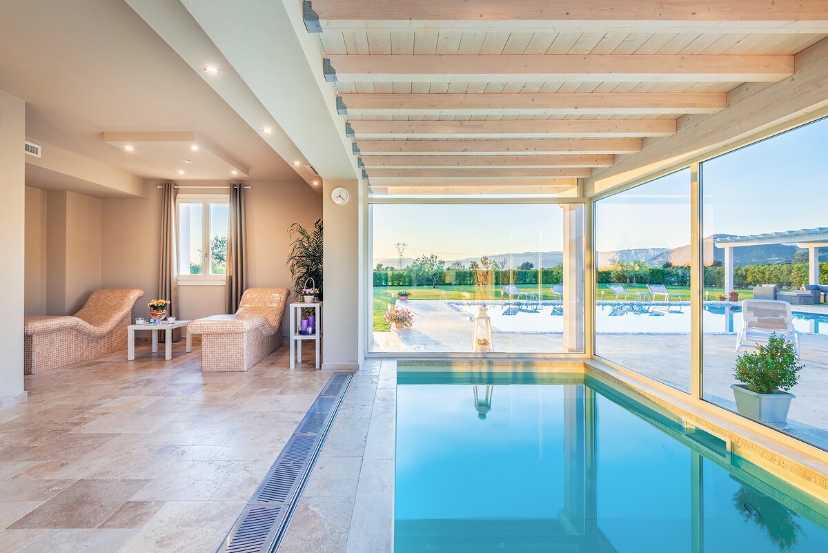 An indoor pool area offers serene views through expansive glass windows. Comfortable seating is arranged nearby, with decorative plants adding a touch of greenery. Natural light brightens the space, highlighting the pool's clear blue water.