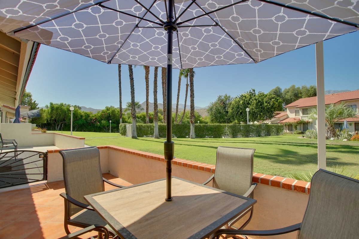 A front patio area featuring a table with an umbrella and four armchairs, surrounded by neatly maintained green lawn and palm trees in the background. The setting offers a view of the community grounds, creating a serene outdoor space for relaxation.