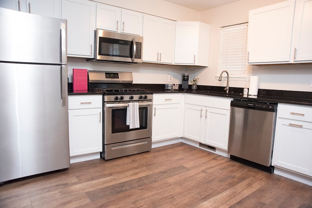 The kitchen features modern stainless steel appliances including a refrigerator, microwave, and oven. White cabinetry offers ample storage, while a black countertop provides a practical workspace. Natural light enters from a nearby window, highlighting the wood-like flooring.