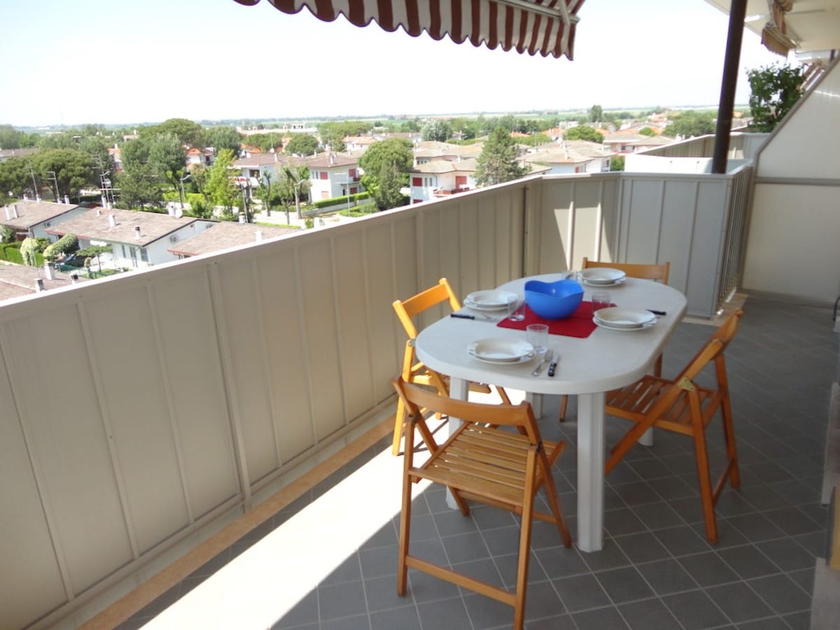 A terrace features a round dining table set with plates and a blue bowl, surrounded by four wooden chairs. The view includes nearby residential buildings and greenery, contributing to a bright and open atmosphere.