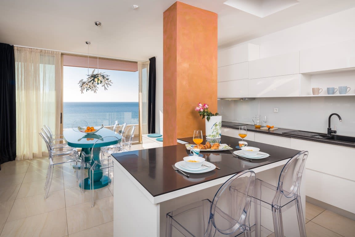 A modern kitchen and dining area is featured, showcasing sleek white cabinetry and a dark countertop. A round glass table is surrounded by transparent chairs, while large windows provide a view of the sea and natural light. Fresh fruit and beverages are arranged on the dining table.