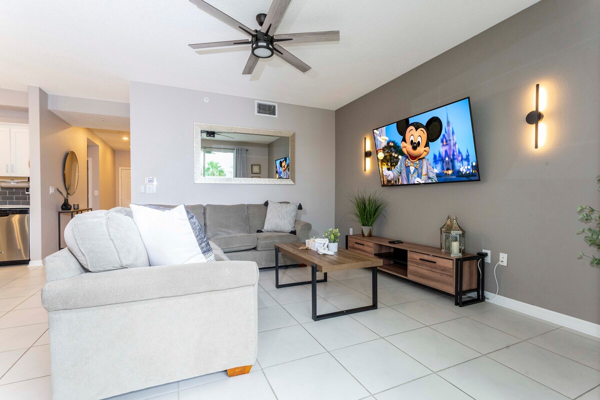 A comfortable living area features a plush sectional sofa in a light beige tone, paired with a wooden coffee table. A large wall-mounted TV displays a Disney character scene. Soft lighting and decorative elements enhance the inviting ambiance of the space.
