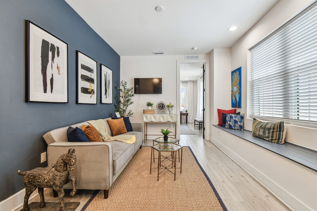 A bright living area is presented featuring a comfortable sofa adorned with colorful cushions. A glass coffee table sits on a natural fiber rug, and artwork lines the walls. A TV hangs on a light-colored wall, while a window seat offers additional seating with decorative pillows.