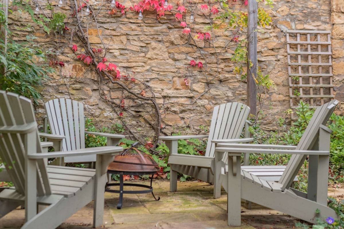 A tranquil outdoor seating area is featured, with four wooden chairs arranged around a circular fire pit. A stone wall adorned with climbing vines provides a natural backdrop, while colorful foliage adds warmth to the inviting space.