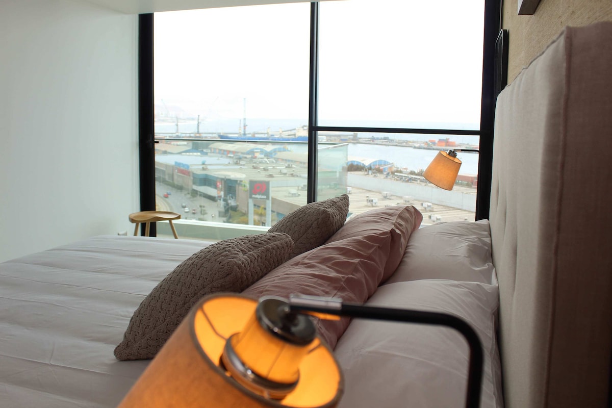 A modern bedroom showcases a spacious bed with neutral-toned linens and two decorative pillows. A bedside lamp is positioned welcomingly, while large windows provide a panoramic view of the harbor and the surrounding urban area.