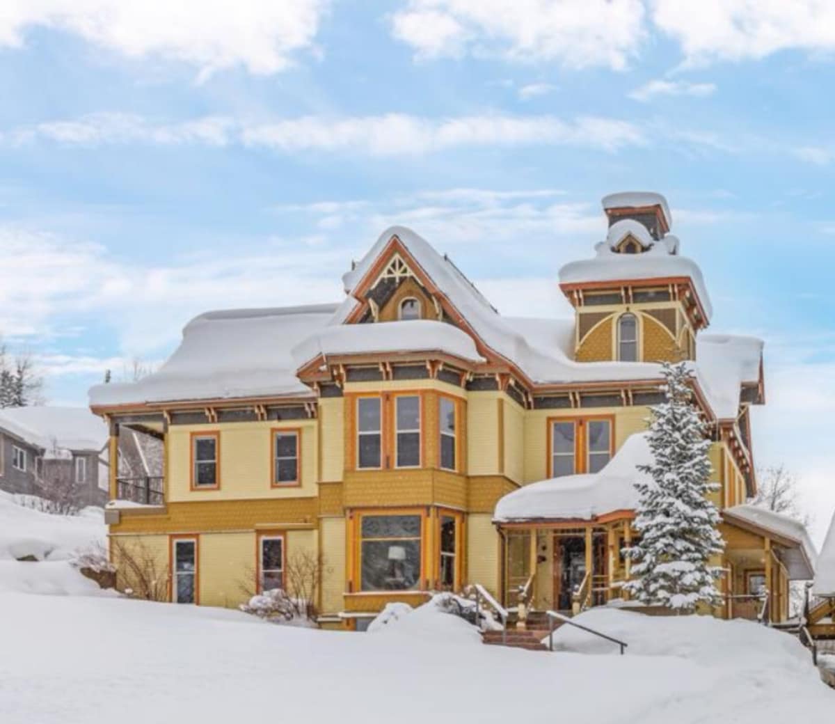 A grand Victorian-style chalet is framed by a blanket of fresh snow, showcasing its intricate architectural details. The yellow exterior stands out against the winter landscape, while a snow-covered roof and surrounding trees add a serene touch to the picturesque setting.