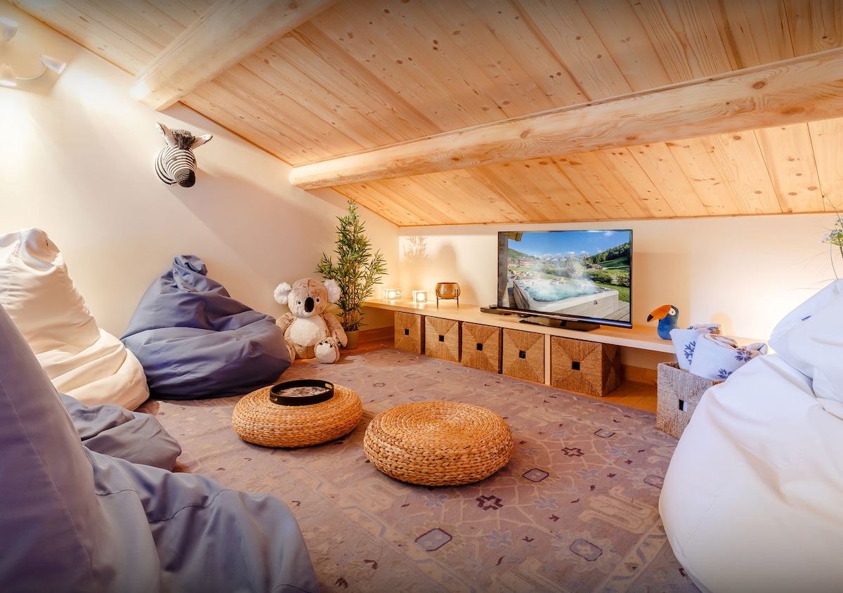 A cozy attic space is furnished with soft bean bags and adorned with decorative pillows. A television is positioned on a low cabinet surrounded by various objects, including plush toys and plants, creating a relaxed atmosphere for entertainment or leisure.