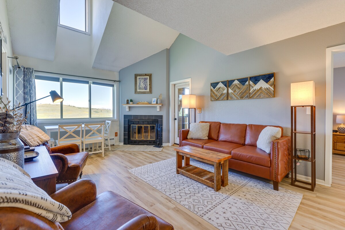 A bright living area features large windows that provide natural light and views of the surroundings. Comfortable seating is arranged around a wooden coffee table, with a wood-burning fireplace as a focal point. Decorative art and a light-colored rug add warmth to the space.
