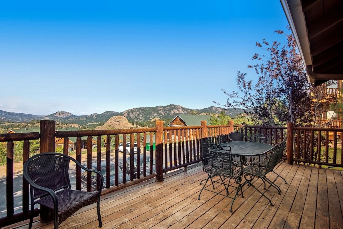 A wooden deck features a table surrounded by chairs, providing a space for outdoor dining. The expansive mountain views in the background create a serene setting, while trees and distant structures add to the natural landscape.