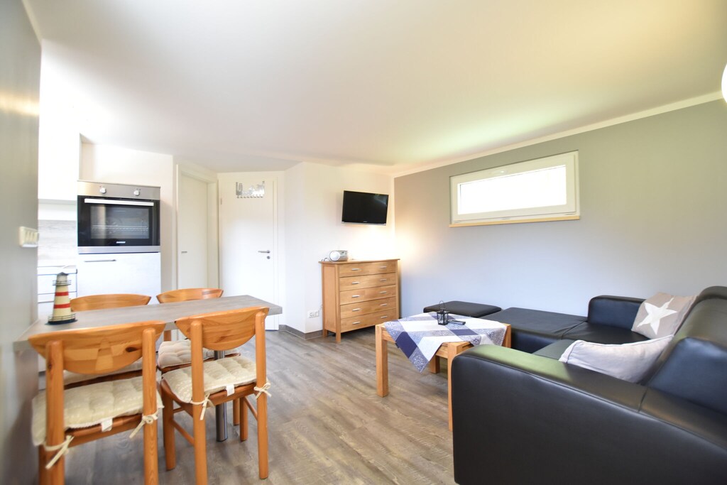 A comfortable living space is furnished with a dark sectional sofa, a light wood dining table with four chairs, and a wooden chest of drawers. A wall-mounted TV is visible, along with modern kitchen elements in the background. Natural light enters through a window, creating a welcoming environment.